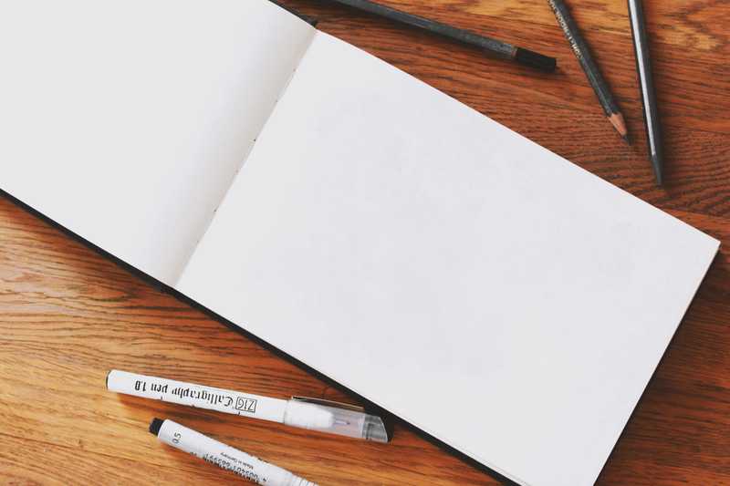 Photo of a blank page in a sketch book with some some pens and pencils next to it.
