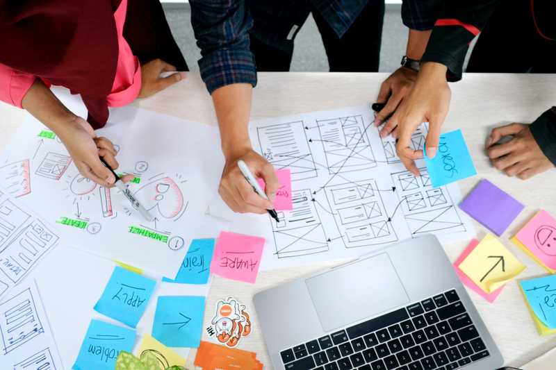 A photo of co-workers brainstorming and solutioning to solve a problem. They are drawing on paper and sticky notes. Key words in the image include 