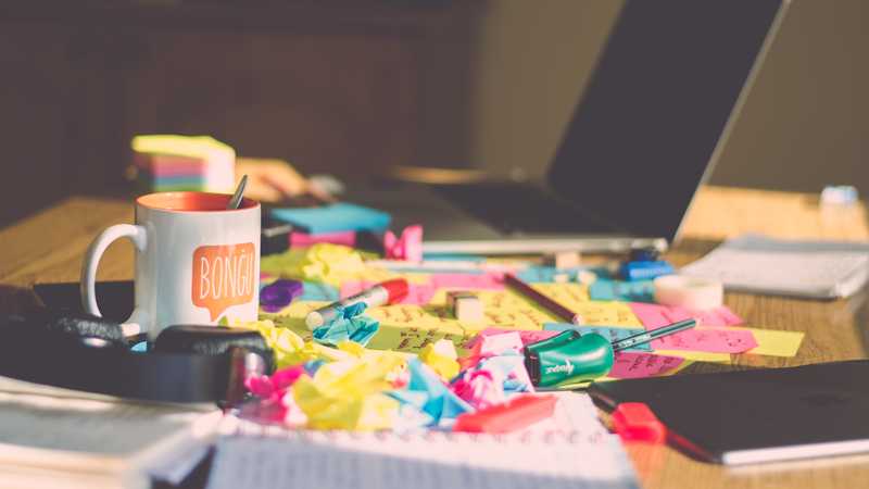 Photo of a messy desk with a computer, coffee cup, crumpled papers, and other office items.